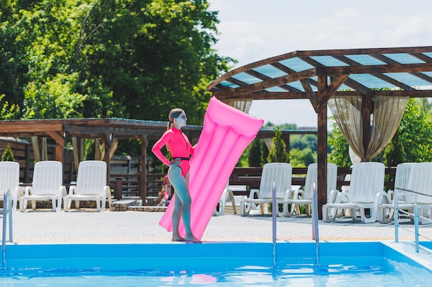 Una bella donna in costume da bagno rosa e con un materasso gonfiabile si trova vicino a una grande piscina Vacanze estive in un hotel con piscina Ragazza in costume da bagno e occhiali da sole