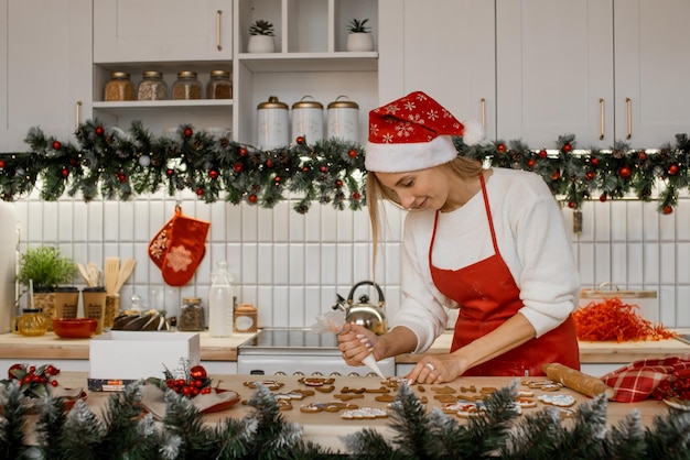 Una bella donna in grembiule rosso e cappello da Babbo Natale sta decorando il pan di zenzero con zucchero a velo nella cucina di Natale
