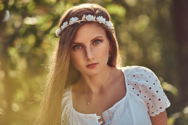 Una bella donna in un vestito bianco e una corona bianca sulla testa che posano in una foresta autunnale verde.