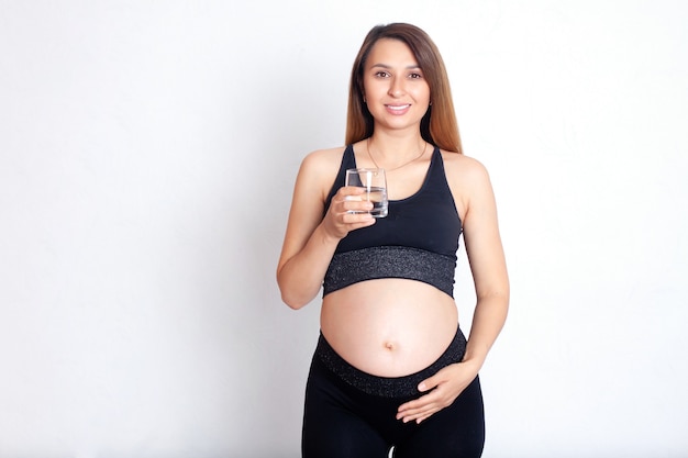 Una bella donna incinta in uniforme sportiva tiene in mano un bicchiere di acqua pulita