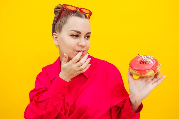 Una bella donna incinta tiene in mano una ciambella dolce su uno sfondo giallo In attesa di un bambino gravidanza e maternità Il concetto di dieta alimentare sana e malsana Cibo spazzatura