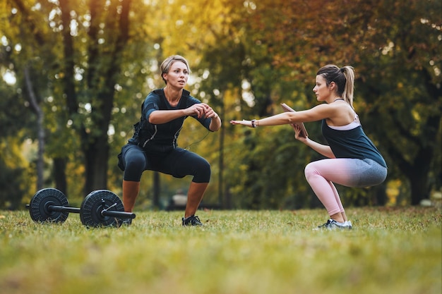 Una bella donna matura sta facendo esercizi di squat con personal trainer nel parco, vestita con un abito nero con un simulatore elettronico EMS per stimolare i suoi muscoli.