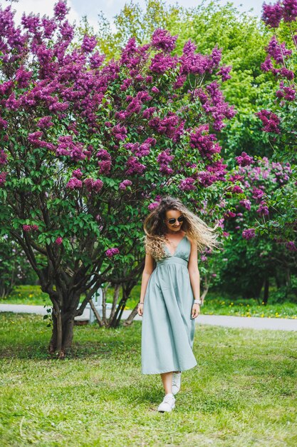 Una bella donna riccia in un abito estivo corre e si rallegra di indossare occhiali da sole sullo sfondo di un cespuglio di lillà viola