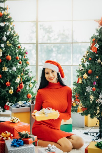 Una bella donna sorridente con un vestito rosso e un cappello da Babbo Natale è seduta circondata da scatole regalo colorate e si gode il Natale da sola a casa.