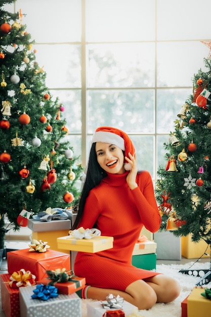 Una bella donna sorridente con un vestito rosso e un cappello da Babbo Natale è seduta circondata da scatole regalo colorate e si gode il Natale da sola a casa.