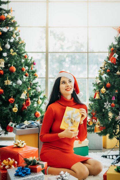 Una bella donna sorridente con un vestito rosso e un cappello da Babbo Natale è seduta circondata da scatole regalo colorate e si gode il Natale da sola a casa.