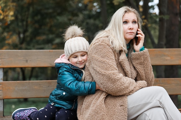 Una bella donna sta parlando al telefono e sua figlia la abbraccia nel parco autunnale