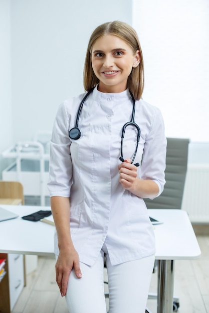 Una bella dottoressa moderna in camice bianco con un fonendoscopio è in piedi in ufficio guardando la telecamera Forma positiva del tirocinante Spazio libero per il testo