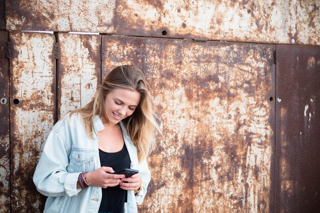 Una bella e bionda donna in una zona urbana che usa il suo telefono sorridente - stile di vita e concetto online e sui social network