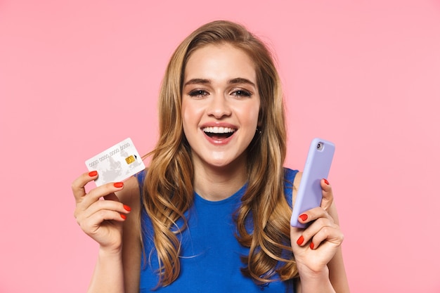una bella e felice giovane donna graziosa in posa isolata su un muro rosa in possesso di carta di credito tramite telefono cellulare