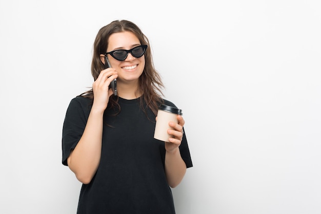 Una bella foto di una giovane donna che beve la sua bevanda calda mentre parla al telefono sorridendo con gli occhiali da sole