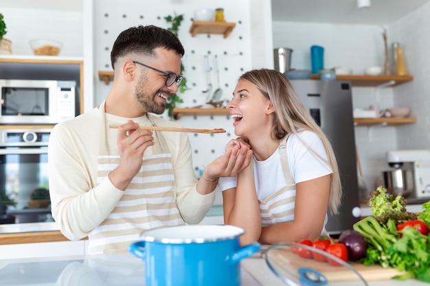 Una bella giovane coppia si guarda e si nutre a vicenda con i sorrisi mentre cucinano in cucina a casa. Una giovane coppia amorevole e gioiosa si abbraccia e cucina insieme divertendosi in cucina.