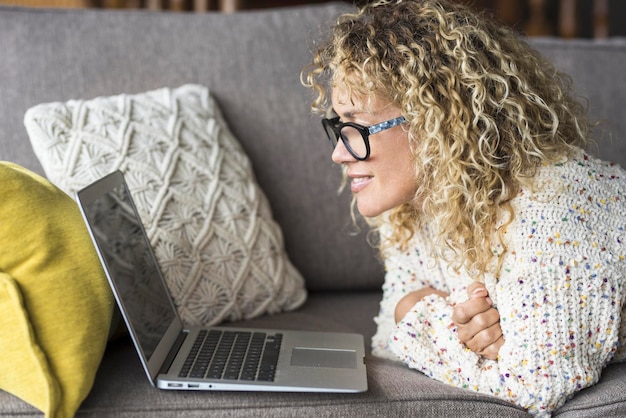 Una bella giovane donna adulta che usa il computer e indossa gli occhiali Persone moderne e connesse che lavorano su un computer portatile sdraiato sul divano di casa Navigando in rete Signora nella rete di videochiamate online Stile di vita su Internet