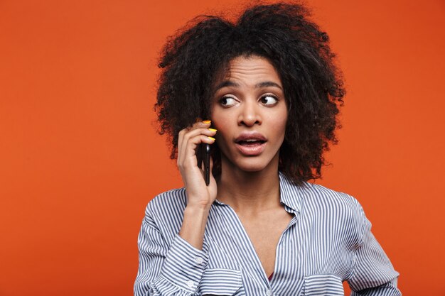 una bella giovane donna africana concentrata isolata sul muro arancione che parla al telefono cellulare.