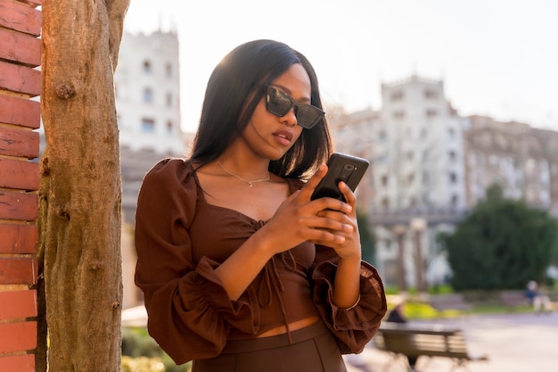 Una bella giovane donna africana naturale in un parco con occhiali da sole guardando il telefono al tramonto
