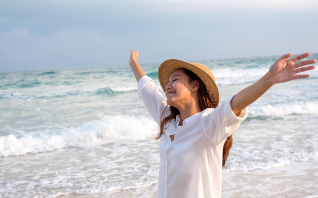 Una bella giovane donna asiatica che alza le mani mentre si passeggia sulla spiaggia con il mare e il cielo blu sullo sfondo