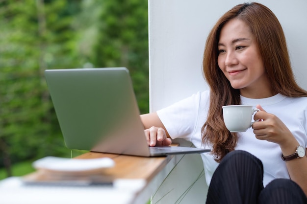 Una bella giovane donna asiatica che beve caffè mentre usa e lavora al computer portatile in mezzo alla natura
