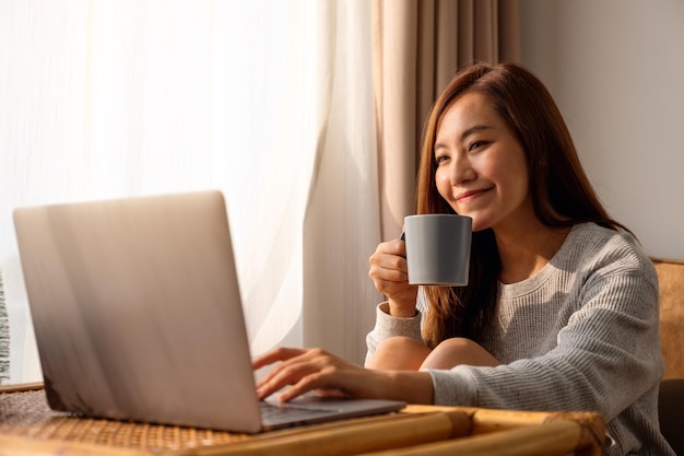 Una bella giovane donna asiatica che usa e lavora al computer portatile mentre beve il caffè a casa