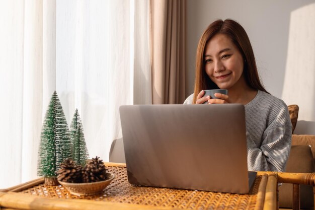 Una bella giovane donna asiatica che usa e lavora al computer portatile mentre beve il caffè a casa