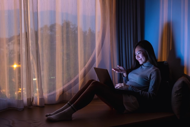 Una bella giovane donna asiatica che utilizza un computer portatile per la videochiamata con schermo luminoso a tarda notte a casa
