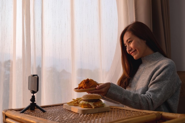 Una bella giovane donna asiatica food blogger o vlogger che mostra e registra un video dal vivo di pollo fritto e hamburger