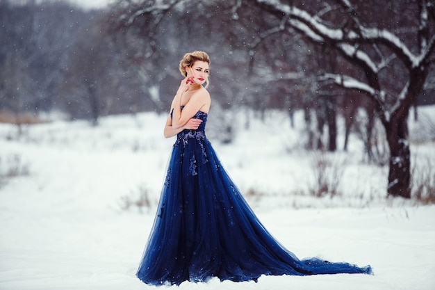 Una bella giovane donna bionda in un abito blu lussureggiante in posa in un parco invernale innevato