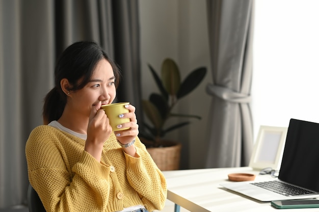 Una bella giovane donna che beve caffè e guarda attraverso la finestra mentre era seduto in ufficio