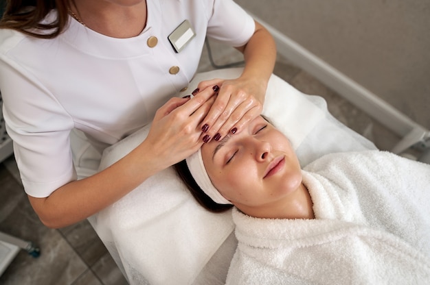 Una bella giovane donna che si distende su un lettino da massaggio mentre lo specialista di bellezza fa un massaggio facciale lifing nel salone SPA