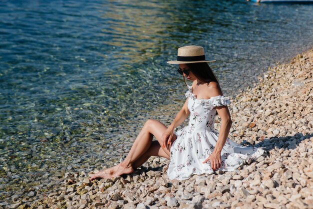 Una bella giovane donna con cappello, occhiali e un vestito leggero è seduta sulla riva dell'oceano sullo sfondo di enormi rocce in una giornata di sole. Turismo e viaggi turistici.