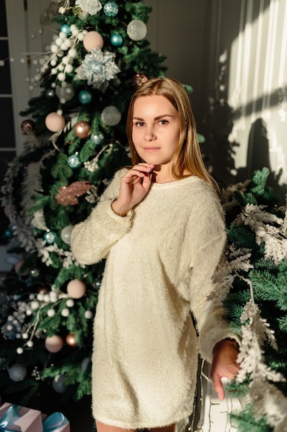 Una bella giovane donna con i capelli biondi si erge sullo sfondo di un albero di Natale. Il nuovo anno sta arrivando presto. Atmosfera natalizia in una casa accogliente