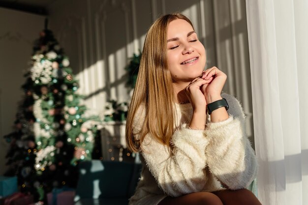 Una bella giovane donna con i capelli biondi si erge sullo sfondo di un albero di Natale. Il nuovo anno sta arrivando presto. Atmosfera natalizia in una casa accogliente