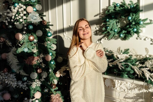 Una bella giovane donna con i capelli biondi si erge sullo sfondo di un albero di Natale. Il nuovo anno sta arrivando presto. Atmosfera natalizia in una casa accogliente