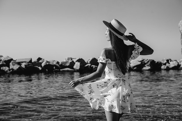 Una bella giovane donna con un cappello e un vestito leggero con la schiena sta camminando lungo la riva dell'oceano sullo sfondo di enormi rocce in una giornata di sole. Turismo e viaggi di vacanza.