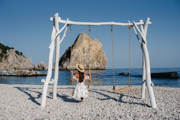 Una bella giovane donna con un cappello e un vestito leggero sta cavalcando un'altalena sulla riva dell'oceano sullo sfondo di enormi rocce. Turismo e viaggi di vacanza.