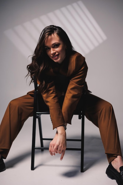 Una bella giovane donna con un sorriso sul viso e indossa un abito classico marrone è seduta su una sedia su uno sfondo grigio Bella donna in una giacca e con il trucco sul viso