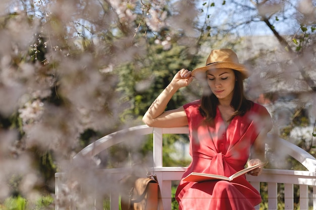 Una bella giovane donna è seduta su un'elegante panchina in un giardino primaverile sotto un fiore di ciliegio e sta leggendo un libro