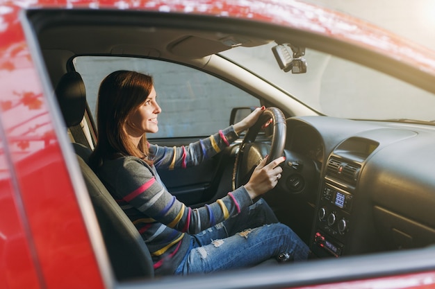 Una bella giovane donna europea dai capelli castani sorridente felice con una pelle sana e pulita vestita con una maglietta a righe si siede nella sua auto rossa con interni neri. Concetto di viaggio e di guida.