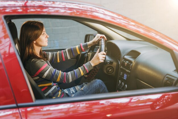 Una bella giovane donna europea dai capelli castani sorridente felice con una pelle sana e pulita vestita con una maglietta a righe si siede nella sua auto rossa con interni neri. Concetto di viaggio e di guida.