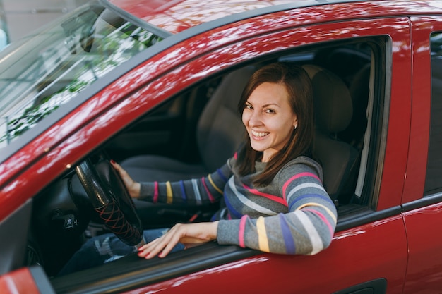 Una bella giovane donna europea dai capelli castani sorridente felice con una pelle sana e pulita vestita con una maglietta a righe si siede nella sua auto rossa con interni neri. Concetto di viaggio e di guida.