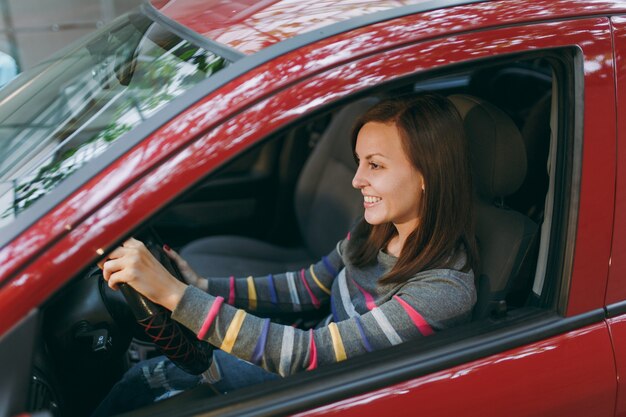 Una bella giovane donna europea dai capelli castani sorridente felice con una pelle sana e pulita vestita con una maglietta a righe si siede nella sua auto rossa con interni neri. Concetto di viaggio e di guida.