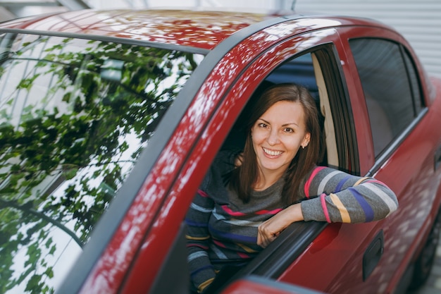 Una bella giovane donna europea dai capelli castani sorridente felice con una pelle sana e pulita vestita con una maglietta a righe si siede nella sua auto rossa con interni neri. Concetto di viaggio e di guida.
