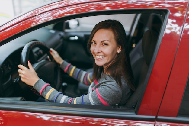 Una bella giovane donna europea dai capelli castani sorridente felice con una pelle sana e pulita vestita con una maglietta a righe si siede nella sua auto rossa con interni neri. Concetto di viaggio e di guida.