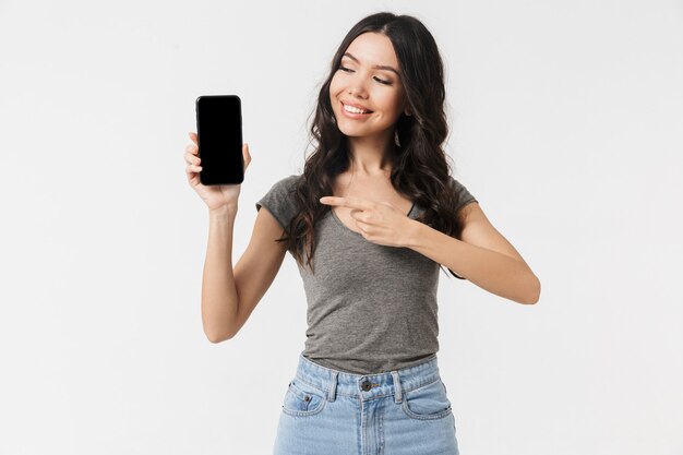 una bella giovane donna felice eccitata in posa isolata sul muro bianco che mostra il display del telefono cellulare.