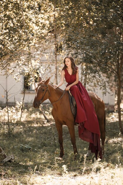 Una bella giovane donna in abito di seta siede a cavalcioni di un cavallo. Passeggiata a cavallo in una giornata di sole nella foresta.