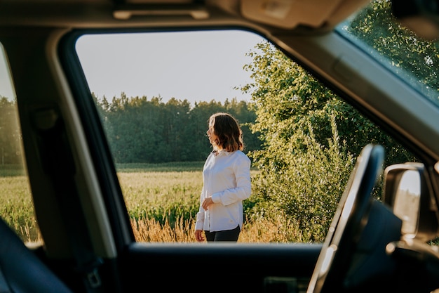 Una bella giovane donna in camicia bianca sta riposando fuori città. All'aperto vicino alla foresta e al campo. Foto attraverso il finestrino dell'auto. viaggio in macchina