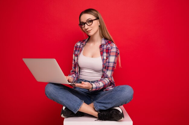 Una bella giovane donna in occhiali e vestiti alla moda è seduta con un laptop su uno sfondo rosso