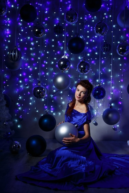 Una bella giovane donna in un lungo vestito blu tiene una palla di natale sullo sfondo di palle e g...