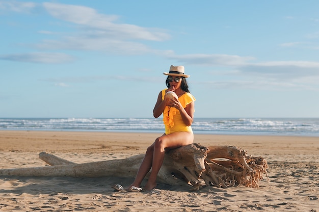 Una bella giovane donna latina in costume da bagno giallo che beve acqua di cocco seduto su un tronco sulla spiaggia