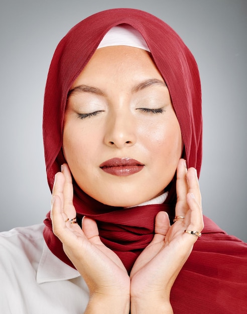 Una bella giovane donna musulmana con gli occhi chiusi che indossa velo rosso e rossetto su sfondo grigio per studio Modesta donna araba che indossa il trucco con la faccia coperta nel tradizionale hijab