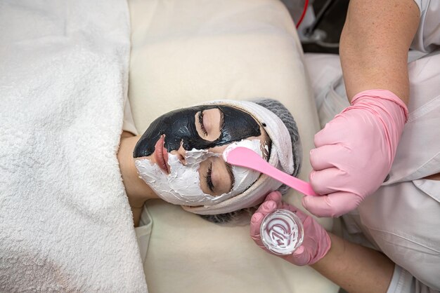 Una bella giovane donna riceve una maschera facciale bianca e nera su metà del viso sdraiata in una spa